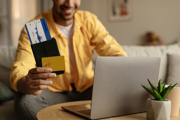 Closeup of young black guy using laptop and booking hotel online with credit card, holding passport...