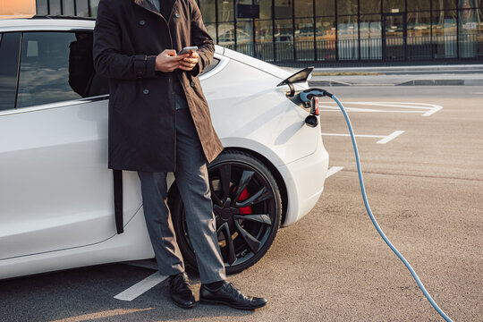 Businessman browsing on a smartphone while waiting to charge his electric car at a charging station in an airport