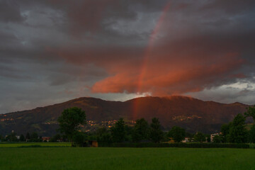 glowing afterglow with rainbow by the sunset in Dornbirn, Vorarlberg, Austria. illuminated red and grey-blue clouds over the green meadows, the city and mountains. god's sign of grace