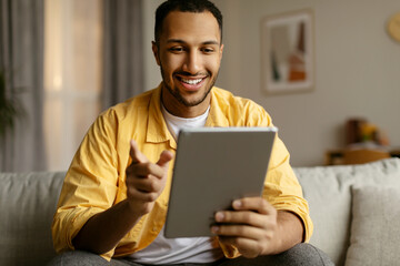 Happy young black man with tablet sitting on couch, working or studying online, having video chat at home