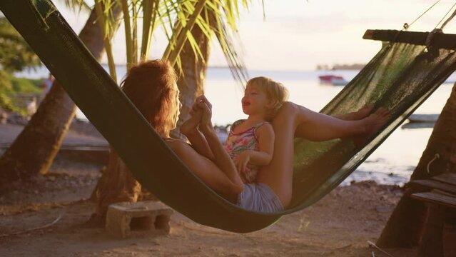 Young woman and her little daughter enjoy a rest in a hammock on the tropical beach to the sound of the surf.
