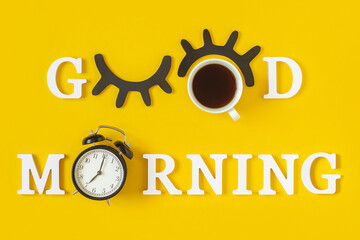 Creative good morning concept. White letters, black eyelashes, alarm clock and cup of coffee on...