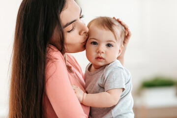 Loving young mother kissing her cute baby, holding little daughter on hands and cuddling, kid looking at camera