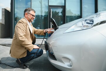 Hansome bearded man sitting near his new modern electric car and holding plug of the charger, while...