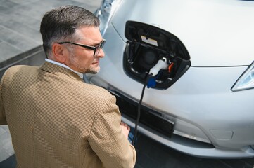 Hansome bearded man sitting near his new modern electric car and holding plug of the charger, while car is charging at the charging station