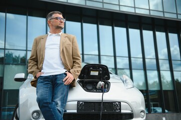 Casual man near electric car waiting for the finish of the battery charging process.