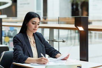 Portrait of young Middle Eastern businesswoman writing in document while working at cafe table in office building setting, copy space