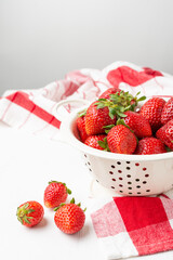 Top view of strawberries in a white colander on white table with red kitchen cloth, white background, vertical