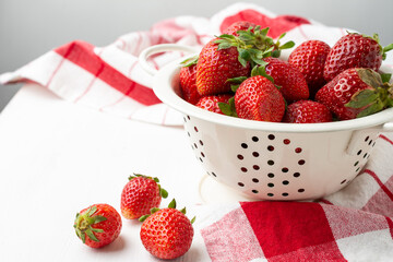 Aerial view of strawberries in a white colander on a white table with a red kitchen towel, white background, horizontal - obrazy, fototapety, plakaty