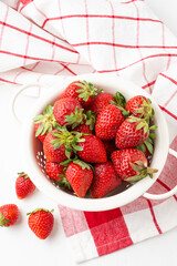 Aerial view of strawberries in a white colander on a white table with a red kitchen cloth, white background, vertical,