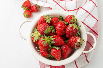 Aerial view of strawberries in a white colander on a white table with a red kitchen cloth, white background with three strawberries, horizontal