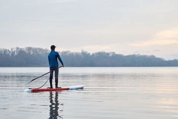 Side view of man on stand up paddle board (SUP) rowing on the river at clear winter day