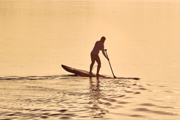 Fototapeta na wymiar Silhouette of woman paddle on stand up paddle board (SUP) on quiet winter or autumn river at sunset. Water sport and meditation on the water
