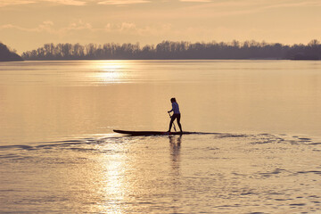 Silhouette of woman paddle on stand up paddle board (SUP) on quiet winter or autumn river at sunset. Water sport and meditation on the water