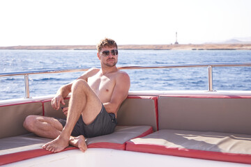 Yacht ride of joyful man sitting on boat and enjoying sea trip.Young man on vacation with sunglasses posing on a yacht and blue sea bay and sky background in a daylight sun.