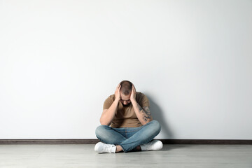 Sad young man sitting on floor near white wall indoors, space for text © New Africa