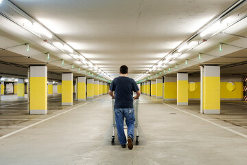 Lonely man walks with a shopping cart in an empty underground parking with yellow concrete columns...