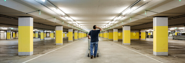 Lonely man walks with a shopping cart in an empty underground parking with yellow concrete columns...