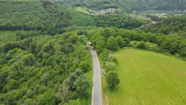 A red bus and three cars travelling along a beautiful road leading down a steep hill in between lush meadows and deciduous forests on a sunny spring morning. Wide angle aerial follow shot