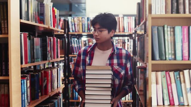 Young male student walks through library among shelves with books holding stack of textbooks in hands, front view. Student holds stack of books in hands