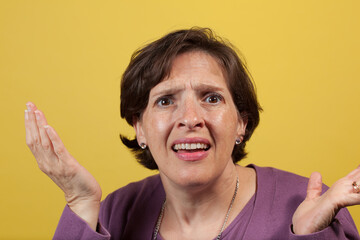 Attractive mature woman in purple blouse on a yellow background with hands apart looking frustrated.