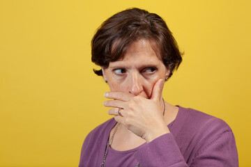 Attractive mature woman in purple blouse on a yellow background with hand over mouth looking away to camera right