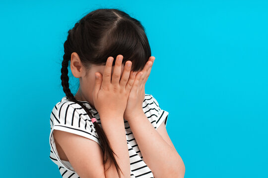 baby girl with pigtails covered her face with her hands on a blue background in a striped dress.