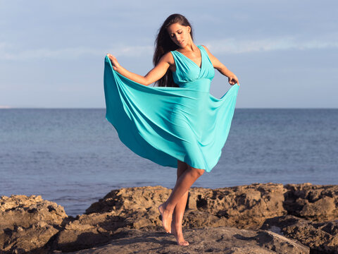 Young Woman In Blue Dress Dancing On The Rocks Beside Sea