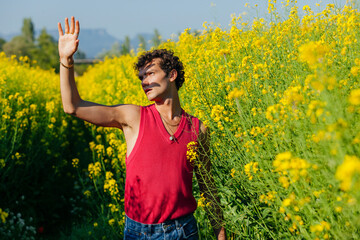 Naklejka na ściany i meble Young homosexual man with curly hair and raised hand, covers his face from the sunlight, standing at yellow oilseed rapes, flower field at spring.