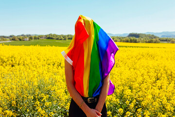 Young gay boy covers his face with rainbow gay pride flag, in yellow flowers field at spring sunny...