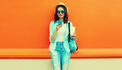 Portrait of happy smiling young woman with smartphone and cup of coffee wearing summer straw hat, backpack on orange background, blank copy space for advertising text