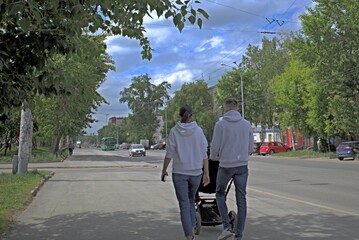 A man and a woman with a baby stroller walk along the sidewalk on a summer day