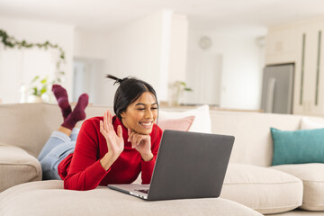Smiling biracial young woman waving hand during video call on laptop at home