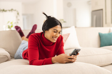 Smiling biracial young woman using smart phone while lying on sofa at home