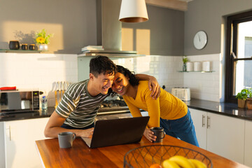 Diverse gay male couple smiling while using laptop together in the kitchen at home