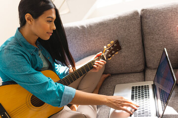 Biracial young woman learning guitar online through laptop at home