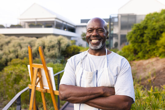 Portrait of smiling bald african american senior man with arms crossed standing by canvas and easel