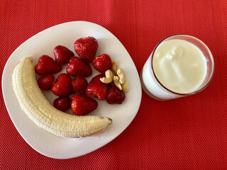 There is a white plate and a glass on a red surface. The plate contains a delicious banana and ripe, red, fresh strawberries and a handful of nuts. White yogurt is poured into a glass.