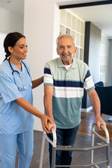 Smiling multiracial female physiotherapist assisting senior man in walking with walker at home