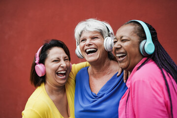 Happy mature female friends having fun together while listeing music with wireless headphone -...