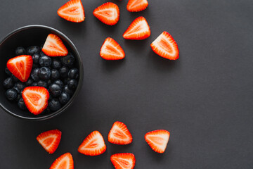 Top view of cut strawberries near blueberries in bowl on black background.