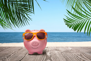 Saving money for summer vacation. Piggy bank with sunglasses on wooden surface near sandy beach and...