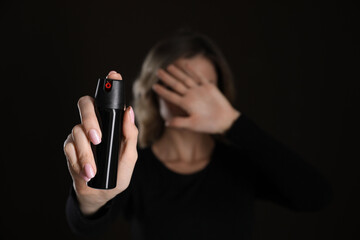 Young woman covering eyes with hand and using pepper spray on black background, focus on canister....