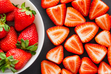 Flat lay with cut and whole strawberries in bowl on black background.