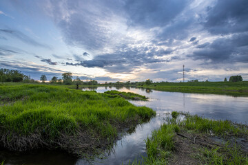 Rural landscape on a spring evening. Dramatic sky reflected in the river. Juicy green grass in the foreground.