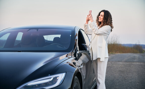 Beautiful curly woman with cute smile standing near her black luxury electric car on road, and taking picture at phone on the backdrop of grey sky.