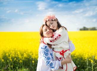 Ukrainian family, mother and daughter, in embroidered shirts, gently hug among blooming yellow...