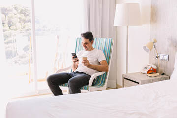 Young man sitting in a rocking chair in his bedroom, looking at the mobile and holding a drink, with the light coming through the window. Concept of working, vacation, break, breakfast, cafe.
