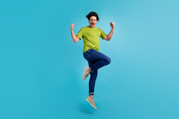 Fototapeta na wymiar Full length photo of funky lucky guy dressed green t-shirt jumping high rising fists isolated blue color background