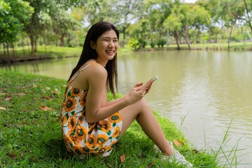 Young beautiful asian woman in sunflower summer dress sitting on the grass and smiling happily while using her phone talking and video calling and enjoying her free time happily in a public park